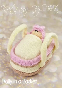 Dolly in a Basket knitting pattern play set – cream-colored knitted basket measuring approximately 18cm long and 11cm wide, containing a 12cm tall pink knitted doll, both crafted with double knitting yarn