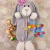 An Easter bunny knitting pattern with a grey body, white paws, and a pink bow on its head. The bunny has a button on its belly and colorful candy-coated chocolates spilling from an opening in its front.