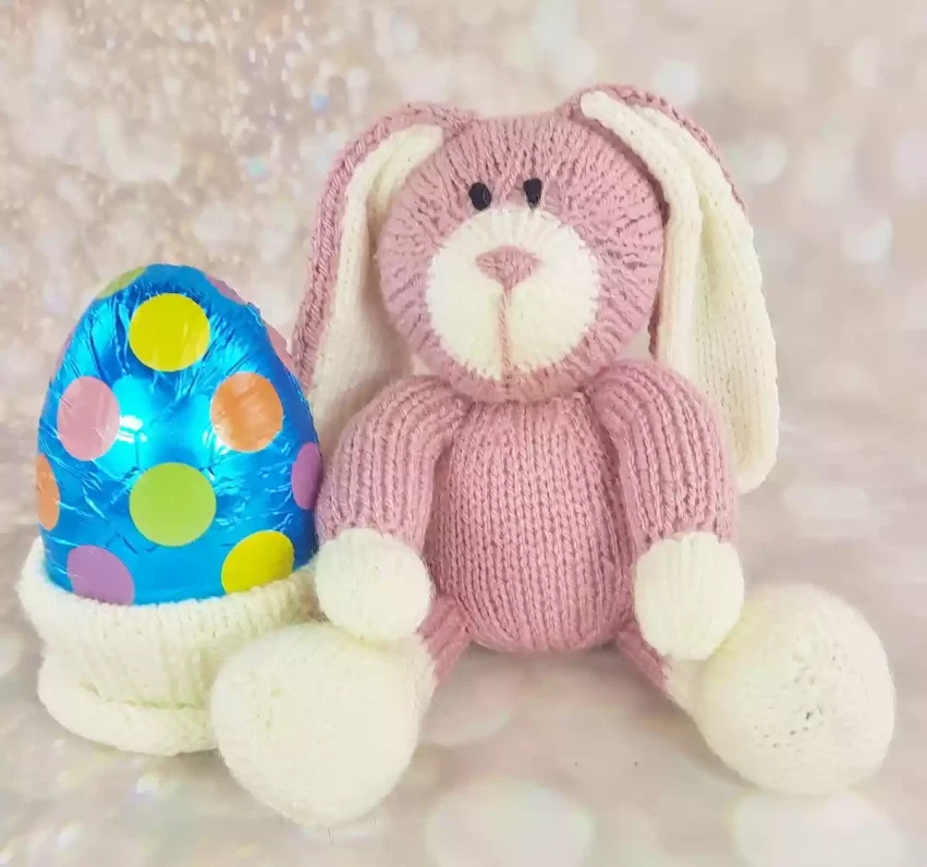 A hand-knitted bunny rabbit with a soft pink body, cream-colored ears, paws, and feet. The bunny has black embroidered eyes, a cream snout, and a pink stitched nose. It is holding a small knitted cream pot containing a large Easter egg wrapped in shiny blue foil with colorful polka dots in orange, yellow, green, and purple.